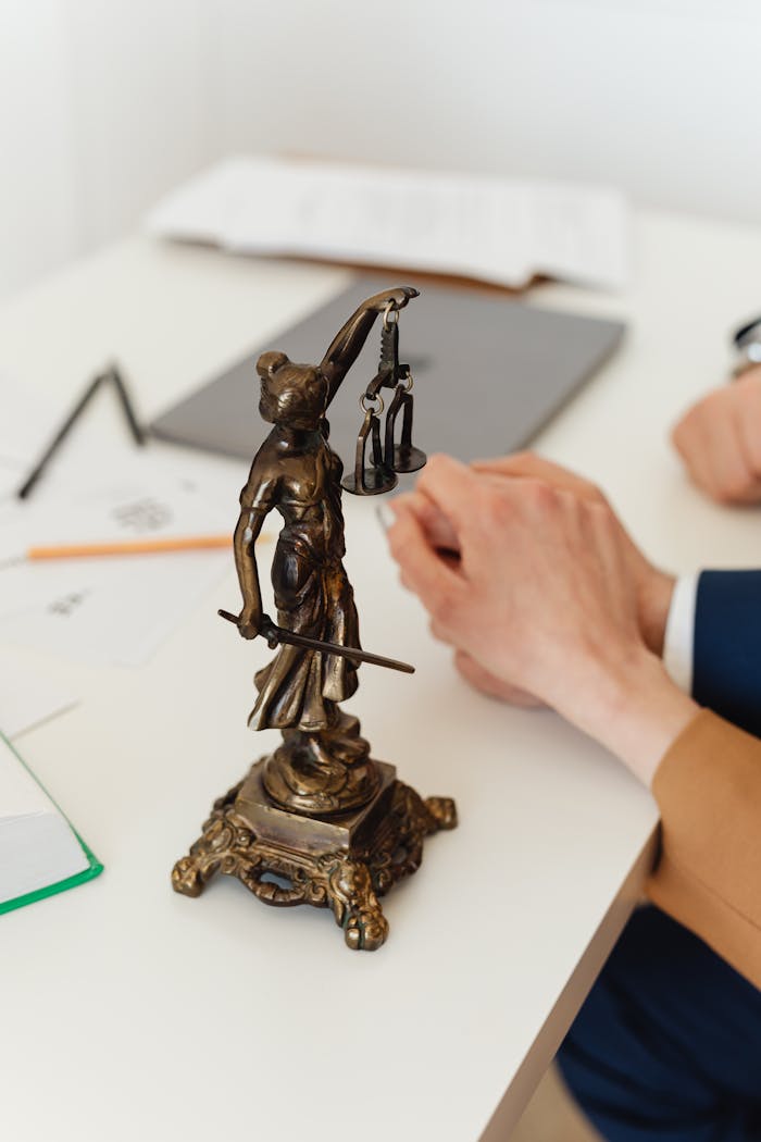 Bronze Lady Justice statue on a white office desk with hands in the background. Symbol of law and fairness.